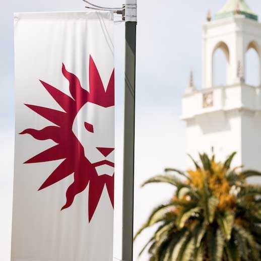 A light pole flag displaying the LMU spirit mark in front of the clock tower