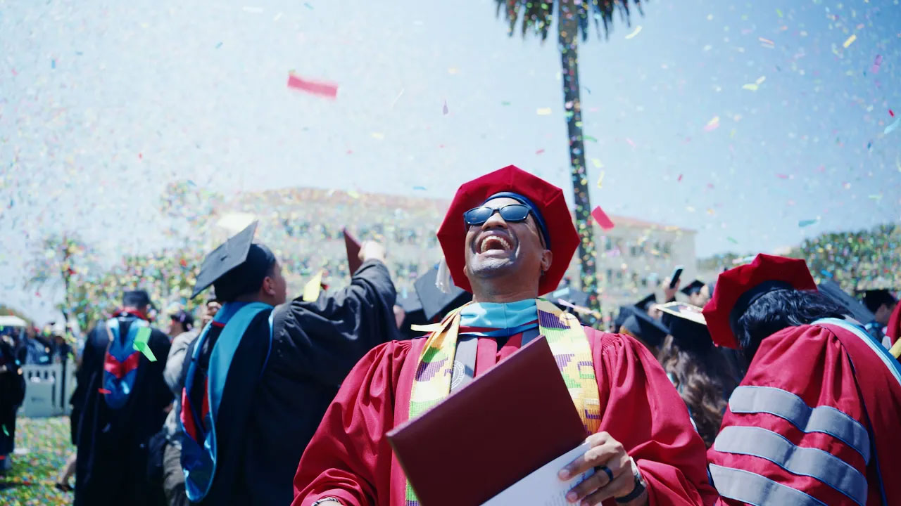 A graduate holding their diploma excitedly looking up at confetti falling around them at the completion of commencement
