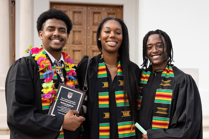 Three graduates wearing their new sashes and standing in front of Sacred Heart Chapel