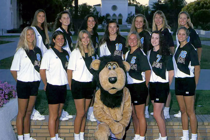 The Womens Volleyball team standing in Alumni Mall with Sacred Heart Chapel, and Iggy kneeling between them