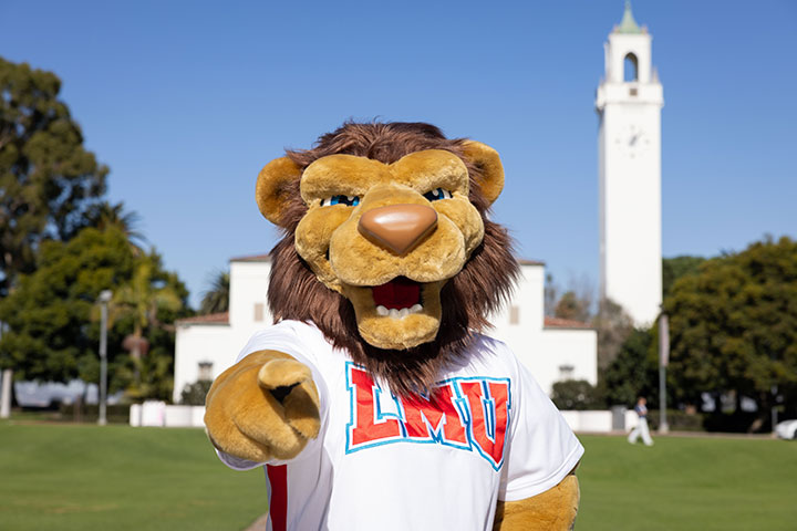 Current mascot design with Iggy standing in front of Sacred Heart Chapel wearing an LMU tshirt and pointing at the camera